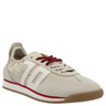 A single beige retro sneaker with white side stripes, a red midsole accent, and red-speckled laces, featuring a suede and nylon upper with a gum rubber sole, presented on a black background.
