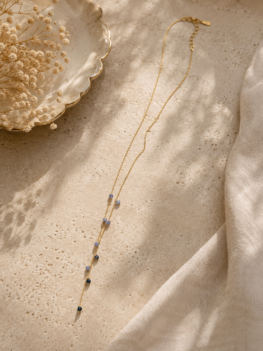 A delicate gold chain necklace with small blue and dark beads, styled on a textured beige surface with soft shadows, draped cream fabric, and dried flowers in a scalloped dish.