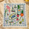 A square embroidered bandana with a colorful Mediterranean-themed design, laid flat on a beige fabric surface with styled props including lemons, a glass, and dried flowers.