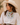 A woman wearing a white cowboy hat, a white blouse, and a white neck scarf with a colorful cocktail and fruit pattern, posed against a warm beige background.
