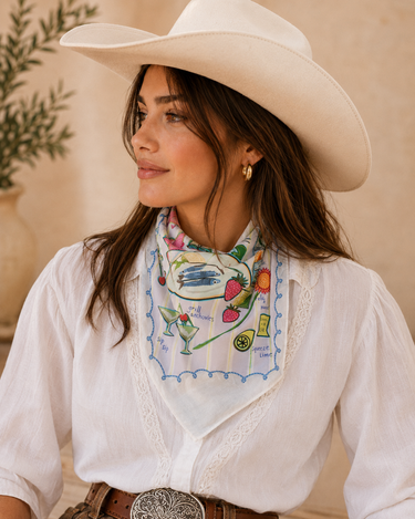 A woman wearing a white cowboy hat, a white blouse, and a white neck scarf with a colorful cocktail and fruit pattern, posed against a warm beige background.