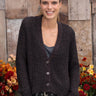 A woman wearing a dark brown chunky knit cardigan with a V-neck and button-front closure, styled over a gray top and jeans, standing in front of a rustic wooden wall with autumn flowers.